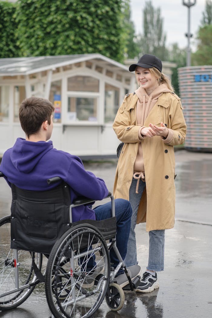 A man in a wheelchair and a woman in a trench coat converse outdoors on a rainy day.